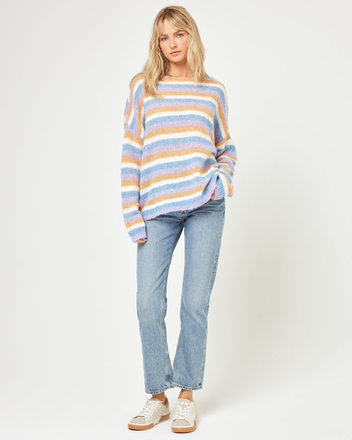Full-length front view of a model wearing the LSPACE Cozy Clouds Sweater in beautiful skies stripe, an oversized fuzzy knit sweater with multicolor horizontal stripes in blue, tan, pink, and white, paired with light jeans and white sneakers on a white background.