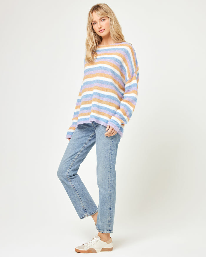 Three-quarter side view of a model wearing the LSPACE Cozy Clouds Sweater in beautiful skies stripe, showing the oversized fuzzy striped knit with multicolor horizontal stripes, styled with light jeans and white sneakers on a white background.