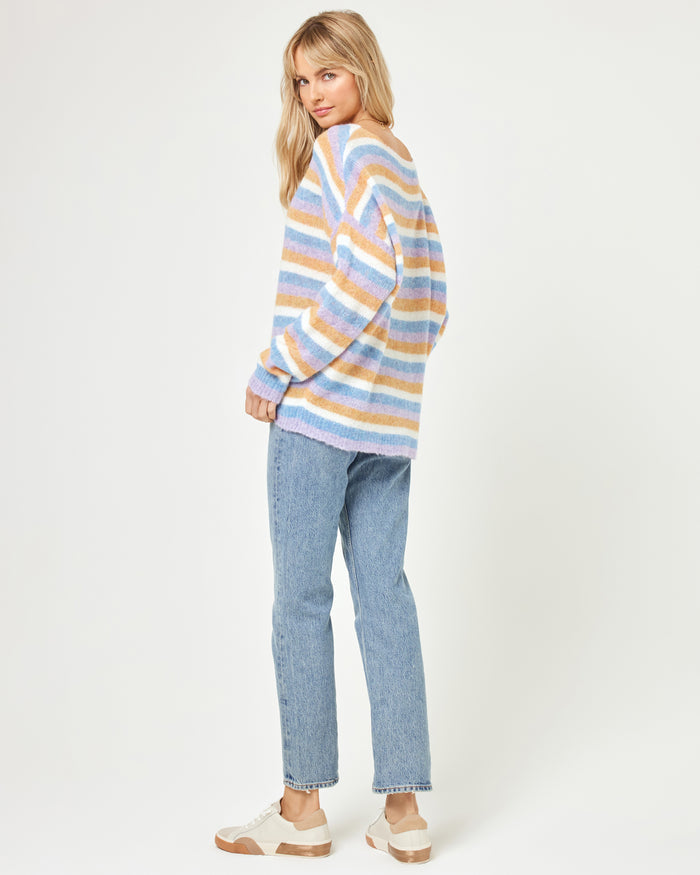 Back three-quarter view of a model wearing the LSPACE Cozy Clouds Sweater in beautiful skies stripe, showing the back of the oversized fuzzy striped sweater in blue, tan, pink, and white, styled with jeans and white sneakers on a white background.