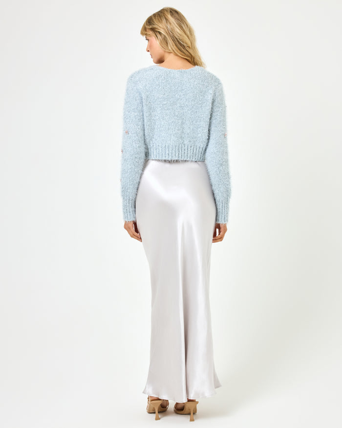 Full body back view of a model wearing the LSPACE Crystal Cardigan in dove, paired with a silver satin maxi skirt, showing the back of the cropped fluffy knit cardigan with long sleeves and ribbed hem.