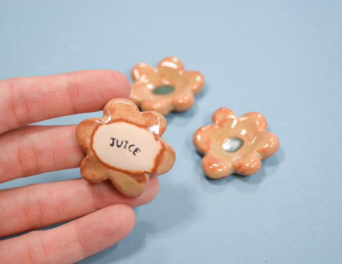 A hand holds a small flower-shaped ceramic cup labeled 'JUICE' with a glossy peach and beige glaze, while two matching cups sit on a light blue surface in the background.