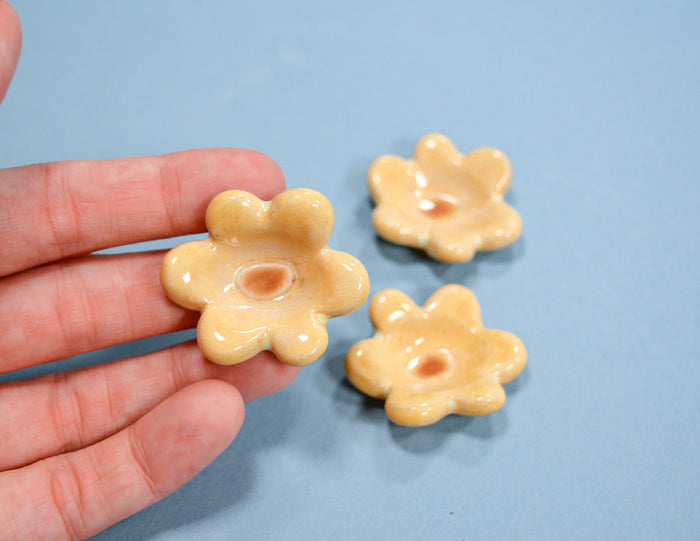 Three small, glossy, flower-shaped beads in a pale yellowish-beige color are shown, with one held in a person's hand and two resting on a light blue background.