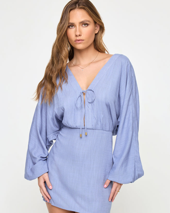 Close-up of a model wearing the LSPACE Deia Mini Dress in chambray, featuring a V-neckline with a tie-front closure and gold hardware, and wide balloon sleeves in chambray blue linen fabric.