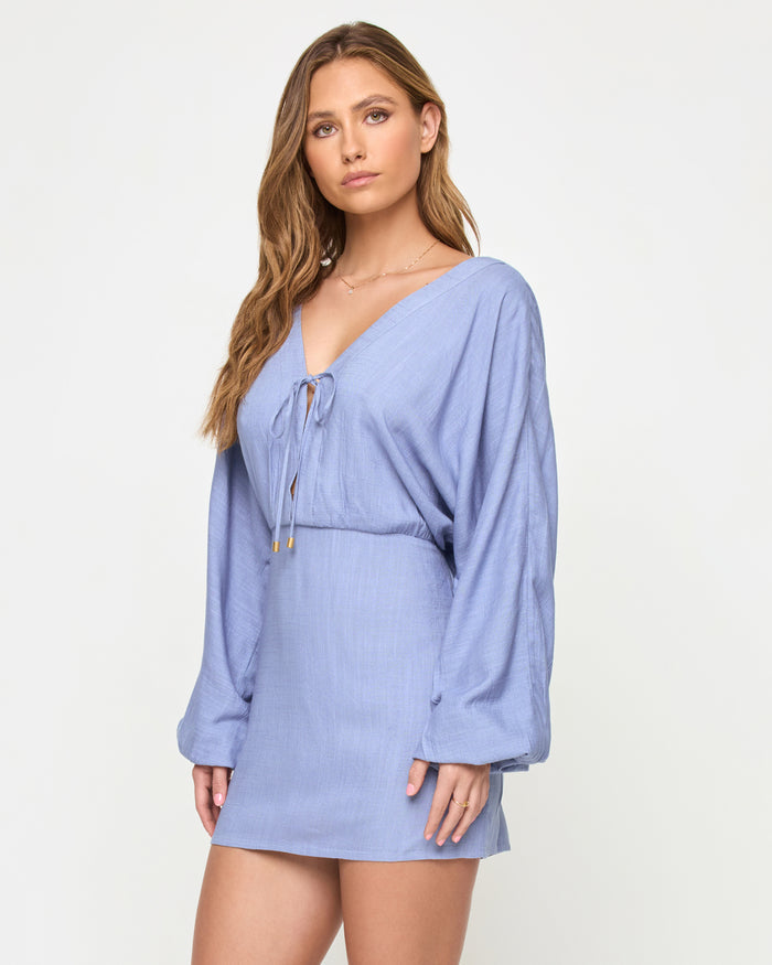 Side view of a model wearing the LSPACE Deia Mini Dress in chambray, showing the profile with wide balloon sleeves, tie-front V-neckline, elastic waistband, and mini length in chambray blue linen fabric.