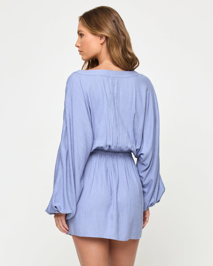 Back view of a model wearing the LSPACE Deia Mini Dress in chambray, showing the open V-back with center seam detail, wide balloon sleeves, elastic waistband, and mini length in chambray blue linen fabric.