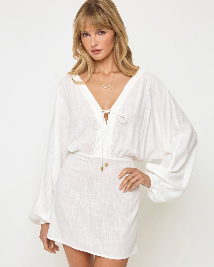 Close-up of a model wearing the LSPACE Deia Mini Dress in cream, showing the deep V-neckline with tie-front closure and gold hardware, wide balloon sleeves, and elasticized waistband in cream textured linen fabric.