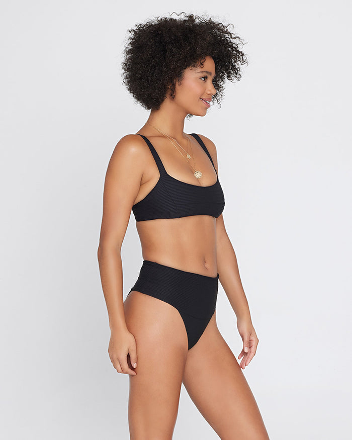 Side view of a model wearing the LSPACE black Desi Bikini Bottom, a ribbed high-waist style, paired with the black Jess Bikini Top.