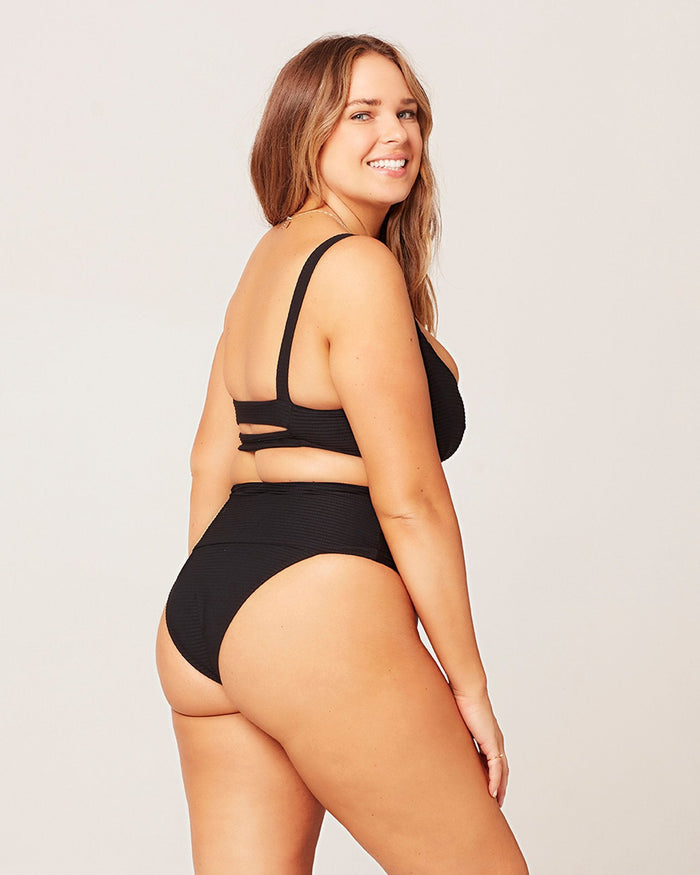 Three-quarter back view of a model wearing the LSPACE black Desi Bikini Bottom, a ribbed high-waist style, paired with a black bikini top with cross-back straps.