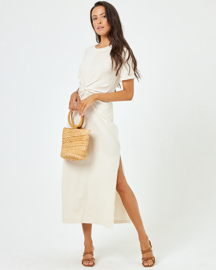 Angled front view of a model wearing the LSPACE Drew Dress in cream, a short-sleeve jersey midi dress with a twisted knot at the waist, styled with a woven tote bag.
