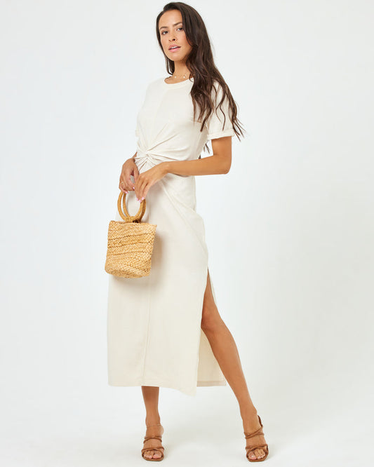Angled front view of a model wearing the LSPACE Drew Dress in cream, a short-sleeve jersey midi dress with a twisted knot at the waist, styled with a woven tote bag.