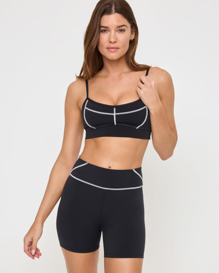Close-up front view from the knees up of the LSPACE Eclipse Bra in black on a model against a white background, showing the bra's trim detail, paired with matching black-cream active shorts.