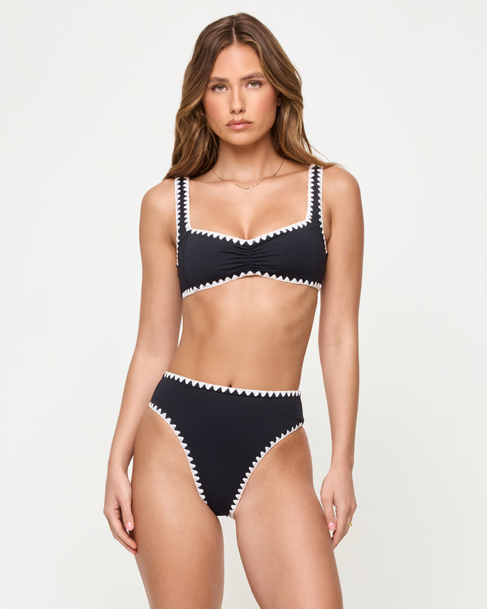 Front view of a model wearing the LSPACE Effie Bikini Top in Black-Cream against a studio background, a black bralette-style bikini top with cream zig-zag trim along the neckline, straps, and hem, paired with matching black high-waist Savannah Bikini Bottom with cream zig-zag trim.