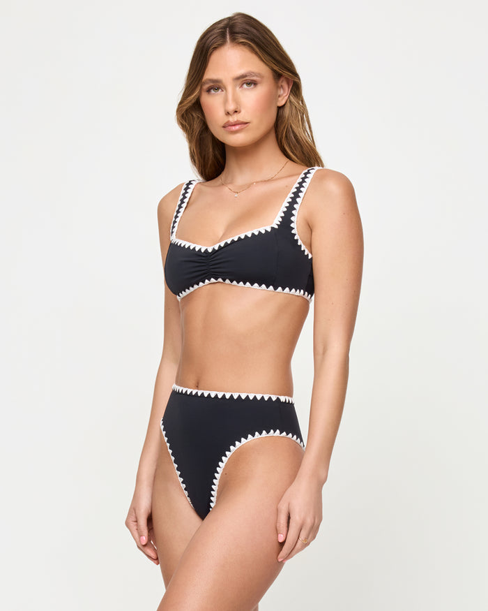 Three-quarter view of a model wearing the LSPACE Effie Bikini Top in Black-Cream against a studio background, a black bralette-style bikini top with cream zig-zag trim along the neckline, straps, and hem, paired with matching black high-waist bikini bottoms with cream zig-zag trim.