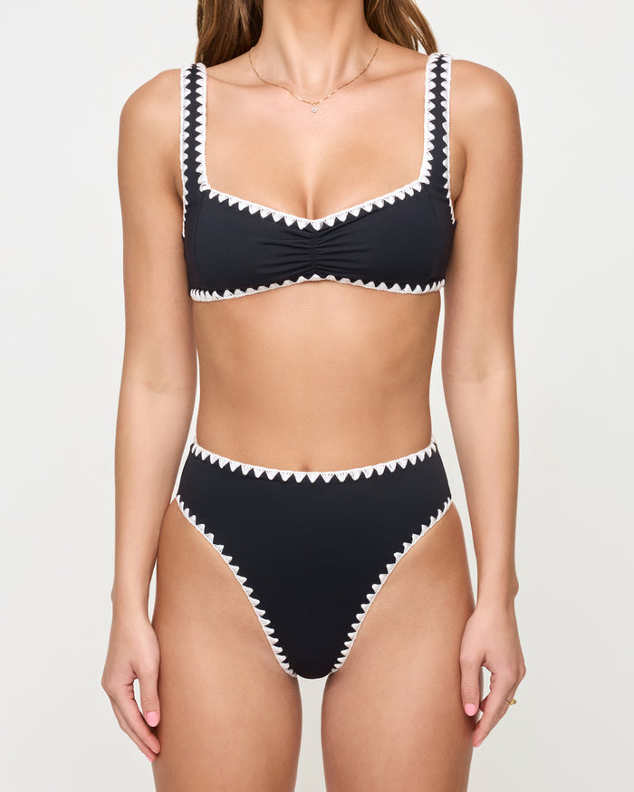 Cropped front view of a model wearing the LSPACE Effie Bikini Top in Black-Cream against a studio background, a black bralette-style bikini top with cream zig-zag trim along the neckline, straps, and hem, paired with matching black high-waist Savannah Bikini Bottom with cream zig-zag trim; model's face is cropped out of frame.