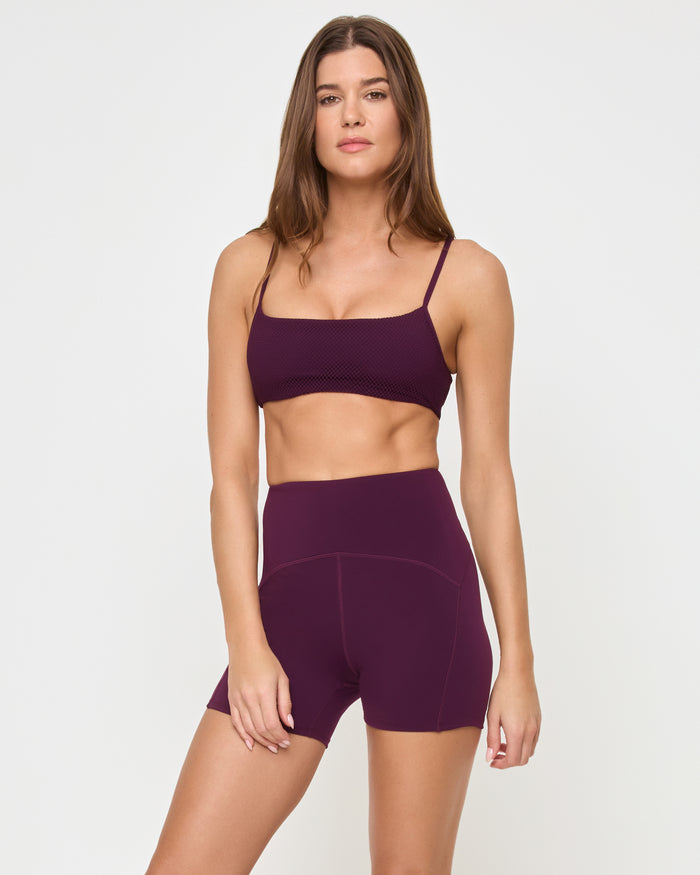 Angled view of the LSPACE En Pointe Short in plum on a model against a white background, showing the profile of the high-waist active shorts, paired with the matching plum Stride Bra.