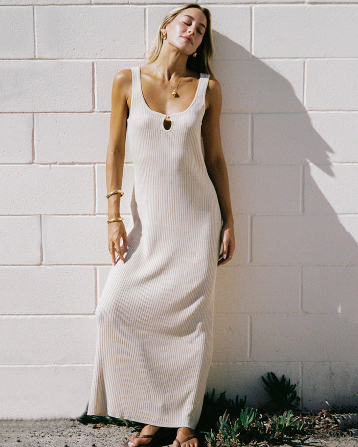 Editorial front view of a model leaning against a white brick wall, wearing the LSPACE En Route Dress - Oat in oat, a ribbed knit sleeveless maxi dress with a keyhole neckline.