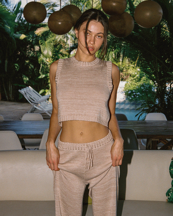 Editorial front view of a model in a tropical indoor setting wearing the LSPACE En Route Top - Heather Oatmeal in heather oatmeal, a sleeveless knit crop top with a crew neckline, styled with matching wide-leg knit pants.