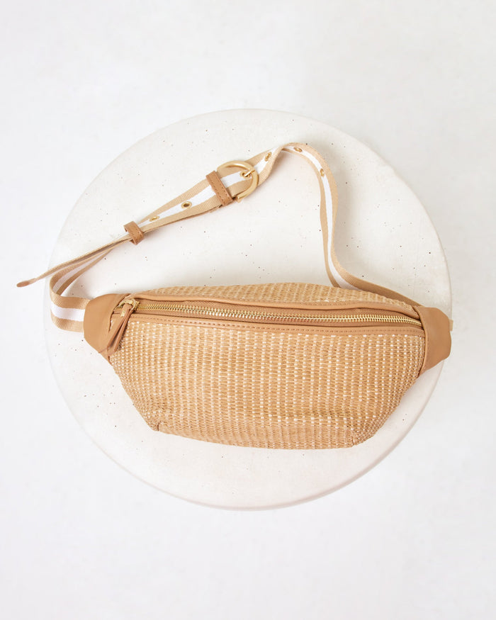 Flat lay of the Evie Fanny Pack in natural on a white pedestal, showing the woven raffia body with tan leather accents and gold zipper.