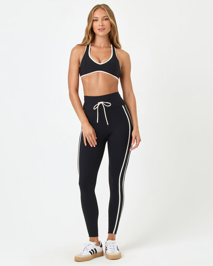Front view of the LSPACE Finley Bra in black-cream on a model against a white background, showing the full halter-style sports bra with cream trim, paired with black high-rise active pants with cream stripe.