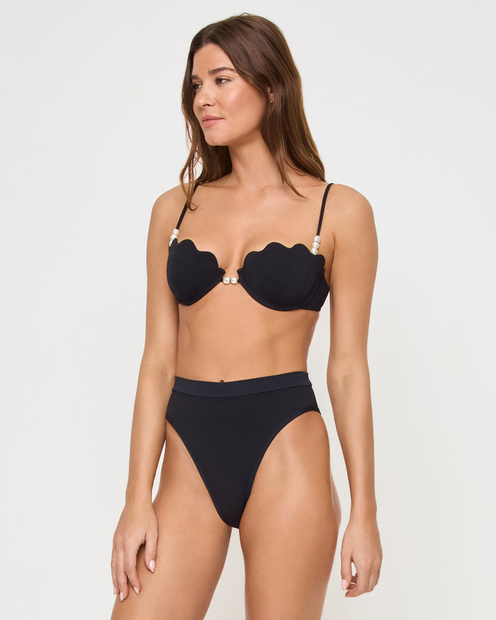 Three-quarter front view of a model wearing the LSPACE black Frenchi Bikini Bottom with a high-waisted style and high-cut leg, paired with a black scalloped Sunrise Shell Bikini Top.