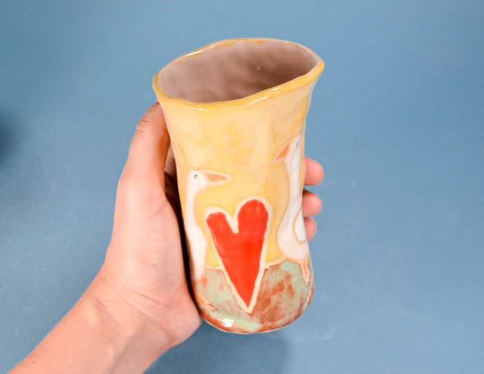 A hand holding a handmade ceramic cup with a yellow and orange glaze, featuring a red heart and white swan design, set against a solid blue background.