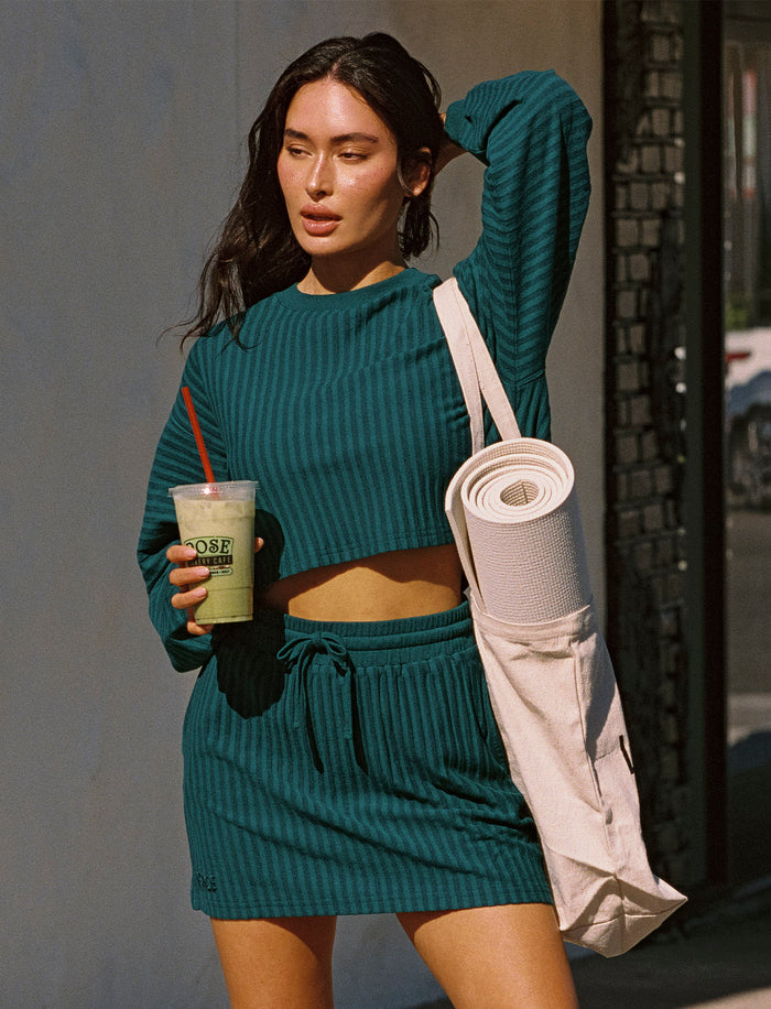 Model outdoors on a city street wearing the Half Court Pullover in deep teal, a ribbed long-sleeve cropped crewneck, with matching deep teal Half Court Skirt, holding a green drink and carrying a yoga mat bag, Adidas sneakers.