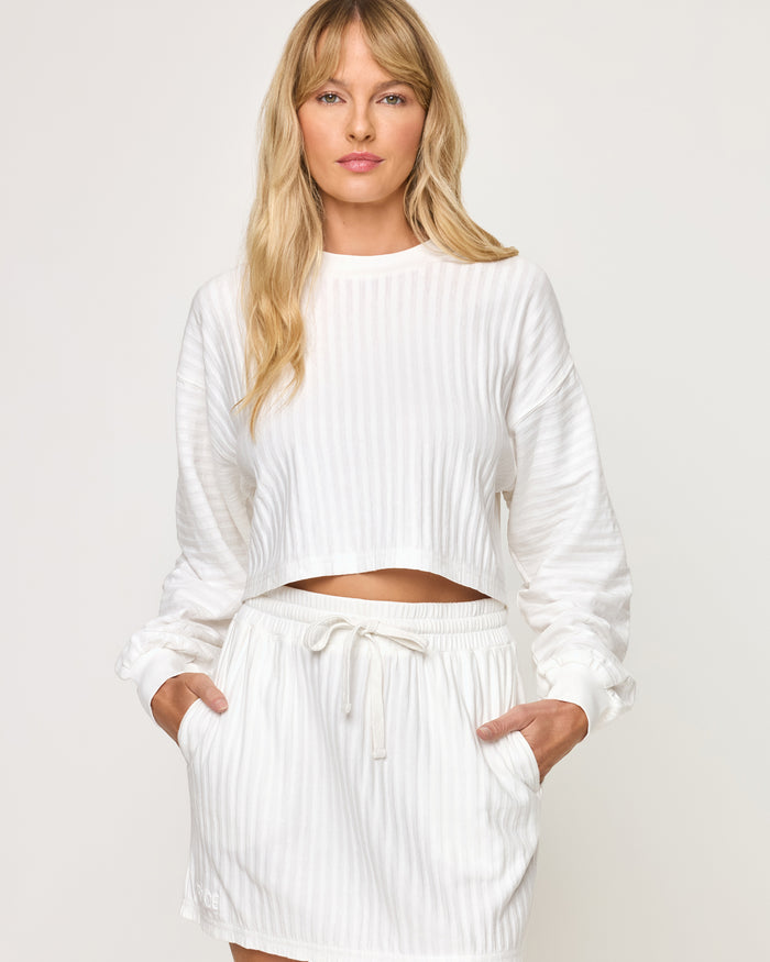 Close-up front view of model wearing the Half Court Pullover in white, a ribbed long-sleeve cropped crewneck, with matching white Half Court Skirt, hands in pockets.