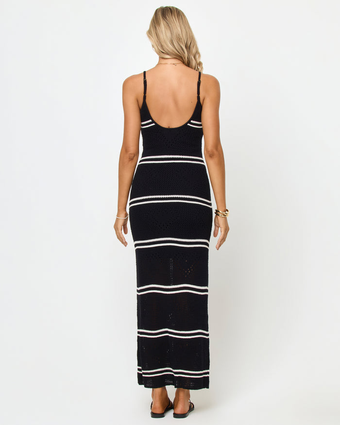 Back view of a model wearing the LSPACE Heart To Heart Dress in black-cream, showing the open low scoop back, spaghetti straps, and body-hugging crochet maxi silhouette in black with horizontal cream stripes.