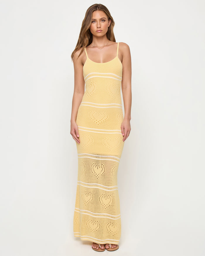 Front view of a model wearing the LSPACE Heart To Heart Dress in daisy-cream, featuring the body-hugging open-knit crochet maxi silhouette in soft yellow with horizontal cream stripe details and heart motifs throughout, scoop neckline, and thin spaghetti straps, against a white studio background.