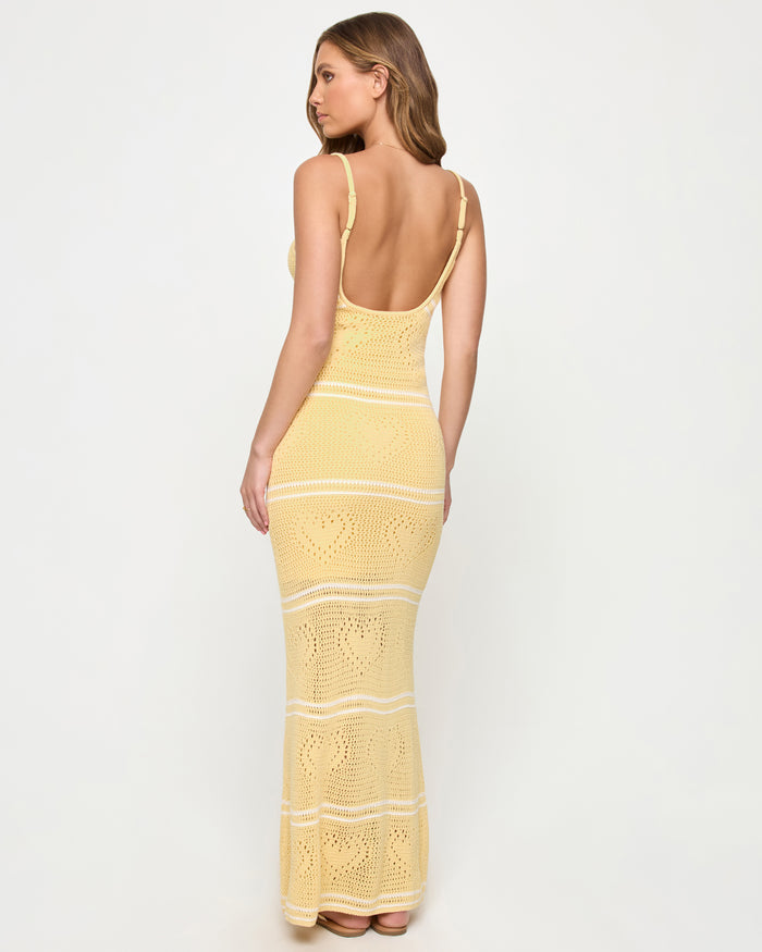 Back view of a model wearing the LSPACE Heart To Heart Dress in daisy-cream, showing the open low scoop back, adjustable spaghetti straps, and body-hugging crochet maxi silhouette in soft yellow with horizontal cream stripe details and heart motifs throughout.