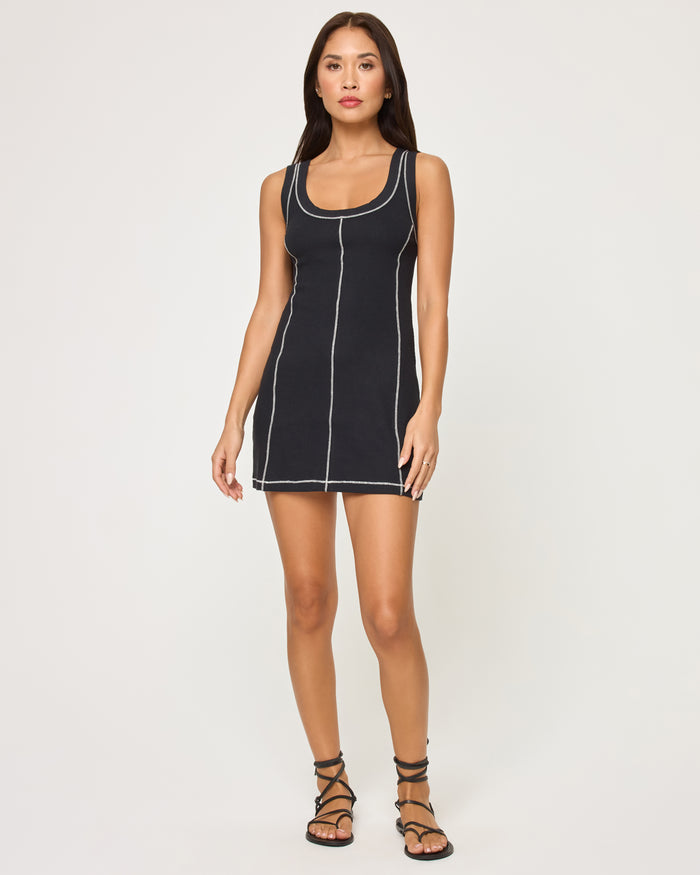 Full-length front view of a model wearing the LSPACE Highline Dress - Black in black, a sleeveless mini dress with contrasting white topstitch seam detailing and a scoop neckline.