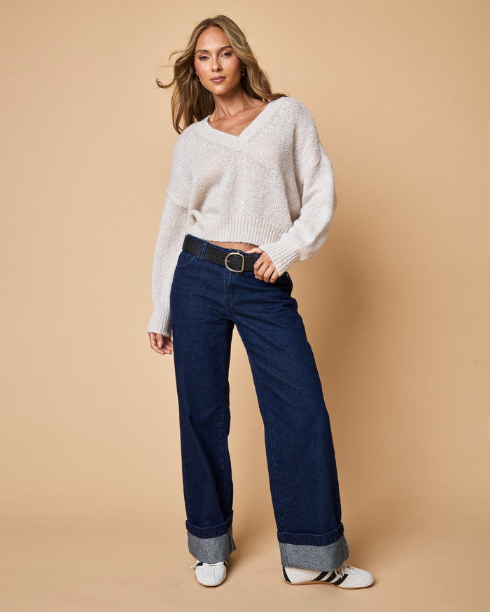 Full-length front view of a model wearing the LSPACE Icon Pullover - Oat in oat, an oversized V-neck knit pullover with long sleeves and chunky knit texture, styled with dark wash wide-leg jeans and sneakers.