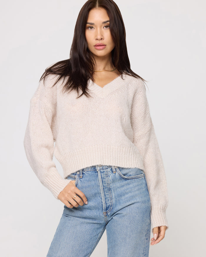 Close-up of a model wearing the LSPACE Icon Pullover - Oat in oat, showing the V-neckline and ribbed hem of the oversized knit pullover, styled with light wash jeans.