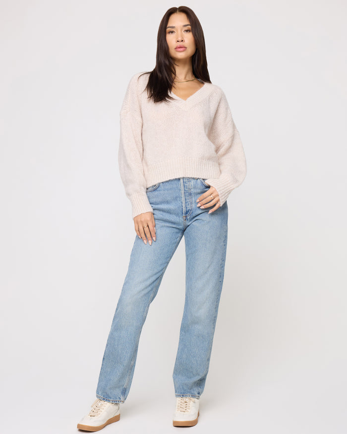 Front view of a model wearing the LSPACE Icon Pullover - Oat in oat, an oversized V-neck knit pullover with long sleeves and ribbed cuffs, styled with light wash jeans and sneakers.