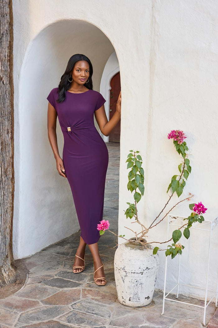 Woman in archway wearing a dark purple short sleeve dress with a gold tone hardware detail on the side