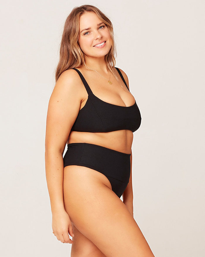 Side view of a model wearing the LSPACE black Jess Bikini Top, a ribbed bralette style with a square neckline, paired with the black Desi Bikini Bottom.
