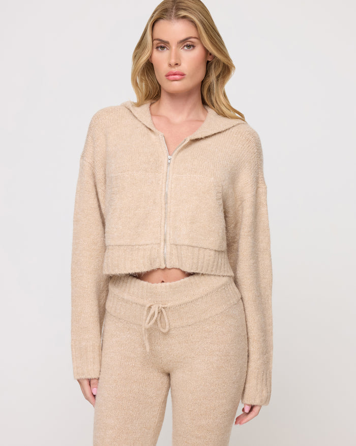 Close-up of a model wearing the LSPACE Jet Set Hoodie - Heather Oatmeal in heather oatmeal, a cropped zip-up hoodie with a ribbed hem and cozy knit texture, styled with matching flared knit pants.