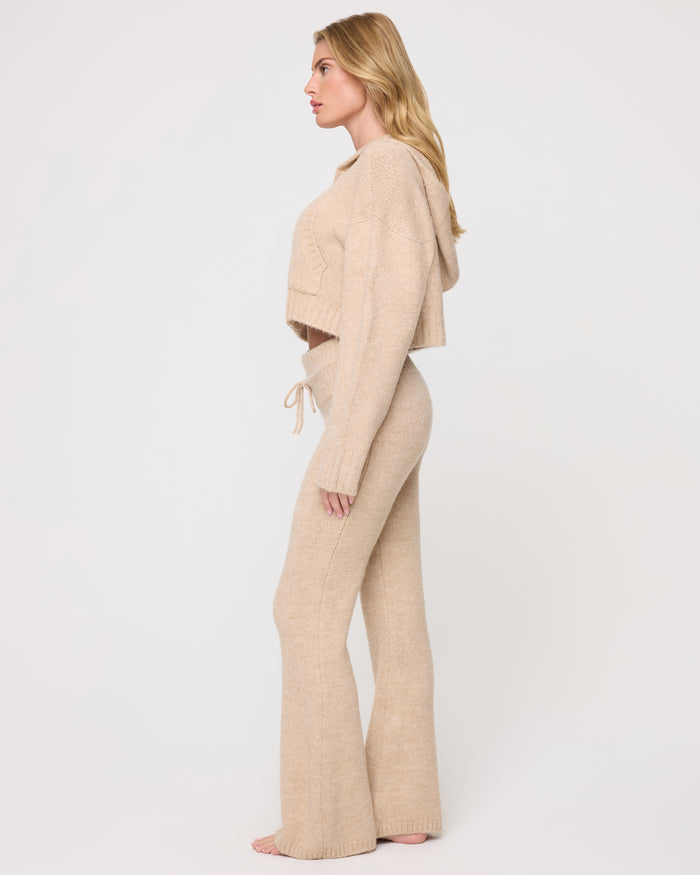 Side view of a model wearing the LSPACE Jet Set Hoodie - Heather Oatmeal in heather oatmeal, showing the cropped length and hood of the zip-up hoodie, styled with matching flared knit pants.