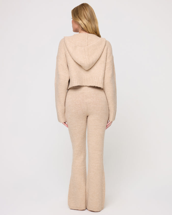 Back view of a model wearing the LSPACE Jet Set Hoodie - Heather Oatmeal in heather oatmeal, showing the back of the cropped zip-up hoodie and the hood, styled with matching flared knit pants.