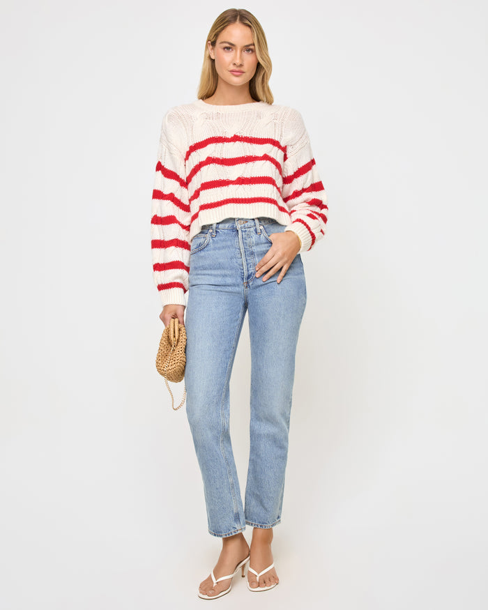 Full-length front view of a model wearing the LSPACE Kensley Pullover in cream flame, a cropped cable-knit sweater with red horizontal stripe detailing and crew neckline, styled with jeans.