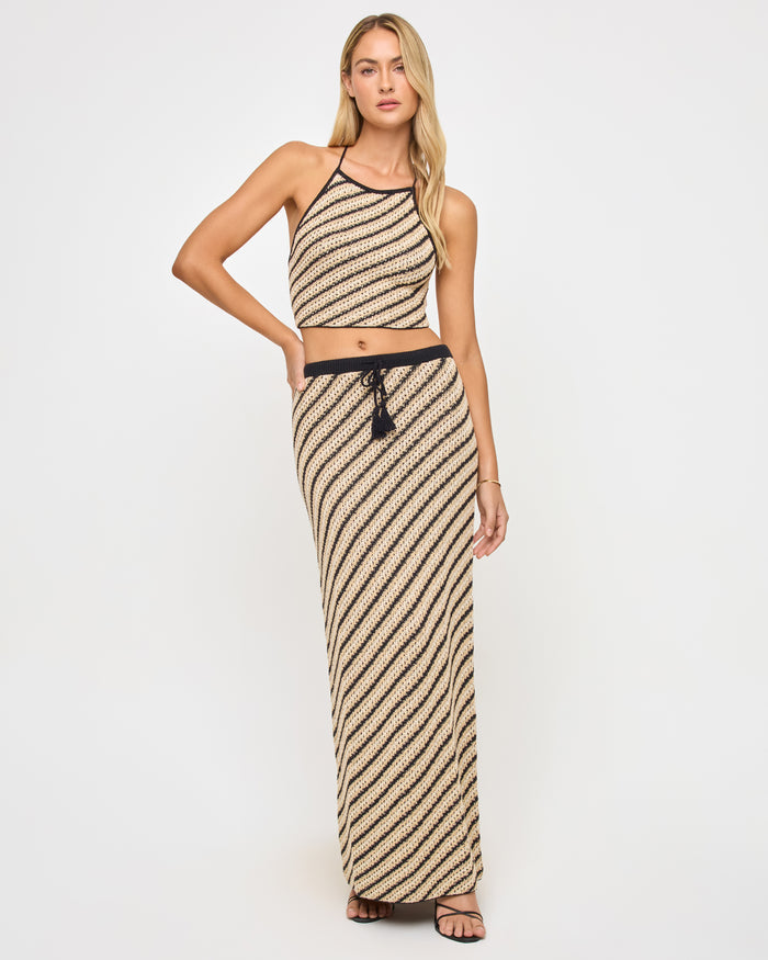 Full-length front view of a model wearing the LSPACE La Bianca Skirt in tip the scales stripe, a maxi-length crochet skirt with diagonal stripe pattern and a black waistband with tassel tie detail.
