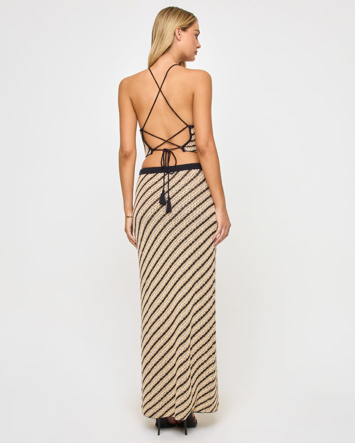 Back view of a model wearing the LSPACE La Bianca Skirt in tip the scales stripe, showing the diagonal stripe crochet pattern and black waistband with tassel tie at the back.
