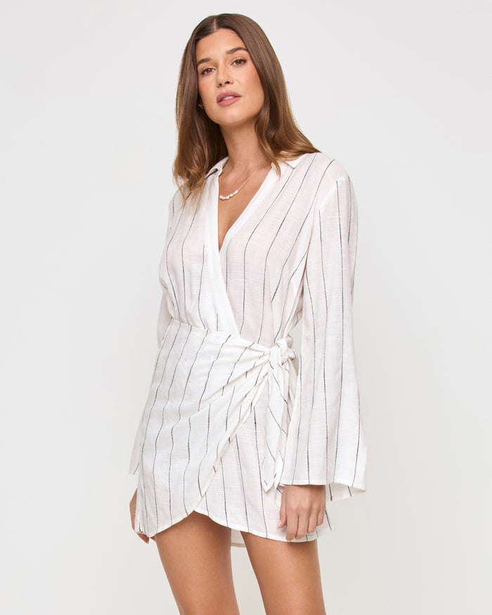 Front view of a model wearing the LSPACE Las Palmas Dress in fine line stripe, a short wrap-style dress with fine vertical stripes, a collared v-neckline, long flared sleeves, and a tie waist.