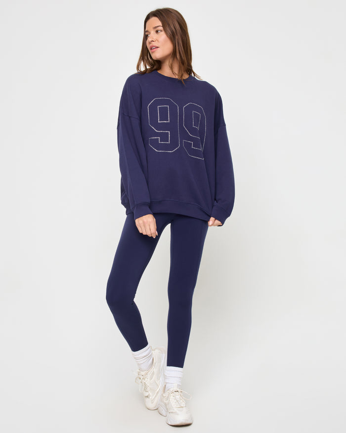 Full-length front view of a model wearing the LSPACE Legend Sweatshirt in midnight blue, an oversized crew-neck sweatshirt with a rhinestone studded '99' graphic, styled with matching navy leggings.