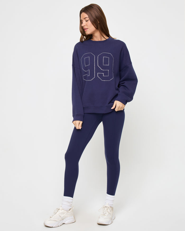 Three-quarter view of a model wearing the LSPACE Legend Sweatshirt in midnight blue, an oversized crew-neck sweatshirt with a rhinestone studded '99' graphic, styled with matching navy leggings and sneakers.