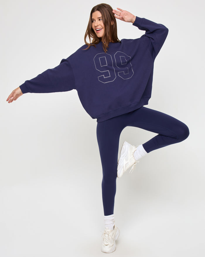 Full-length view of a model wearing the LSPACE Legend Sweatshirt in midnight blue, an oversized crew-neck sweatshirt with a rhinestone studded '99' graphic, shown in a playful pose with matching navy leggings.