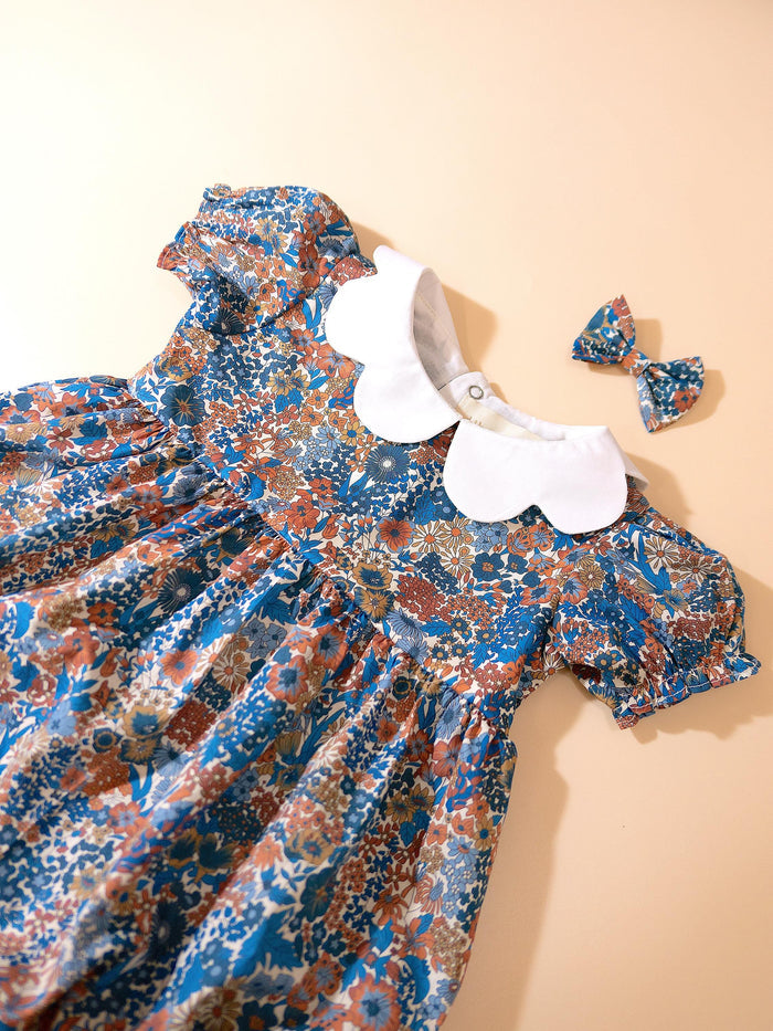 May include: A blue floral print dress with a white Peter Pan collar and matching bow. The dress features short, puffed sleeves and a gathered waist. The floral pattern includes shades of blue, orange, and brown. The dress is designed for a child.