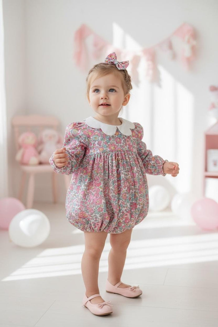 May include: A young child wearing a floral print romper with a white collar and matching bow. The long-sleeved romper features a pink and white floral pattern. The child is also wearing pink ballet flats. The background includes pink balloons and a pink chair.