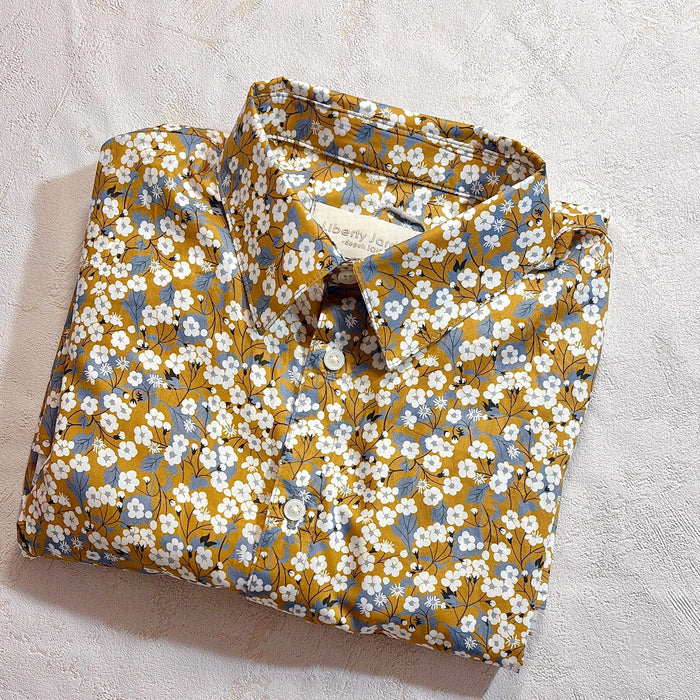 May include: A folded button-down shirt with a floral pattern. The shirt features a mustard yellow background with white and blue floral accents. The collar is visible, and the shirt is neatly folded.