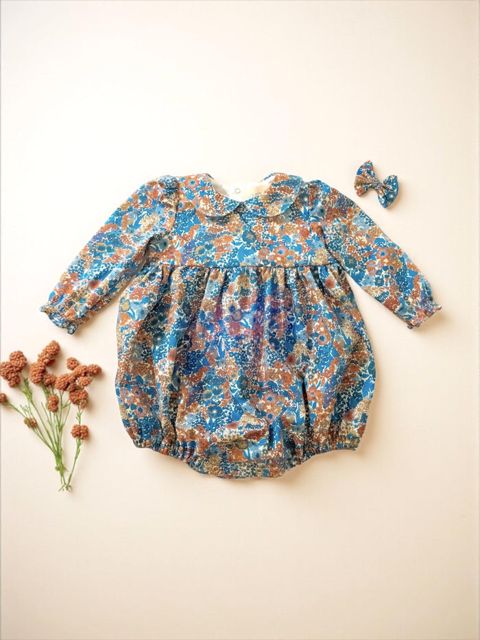 May include: A blue, brown, and white floral print baby romper with long sleeves and a Peter Pan collar. A matching bow hair clip is included. The romper is made of a soft fabric and is designed for comfort.